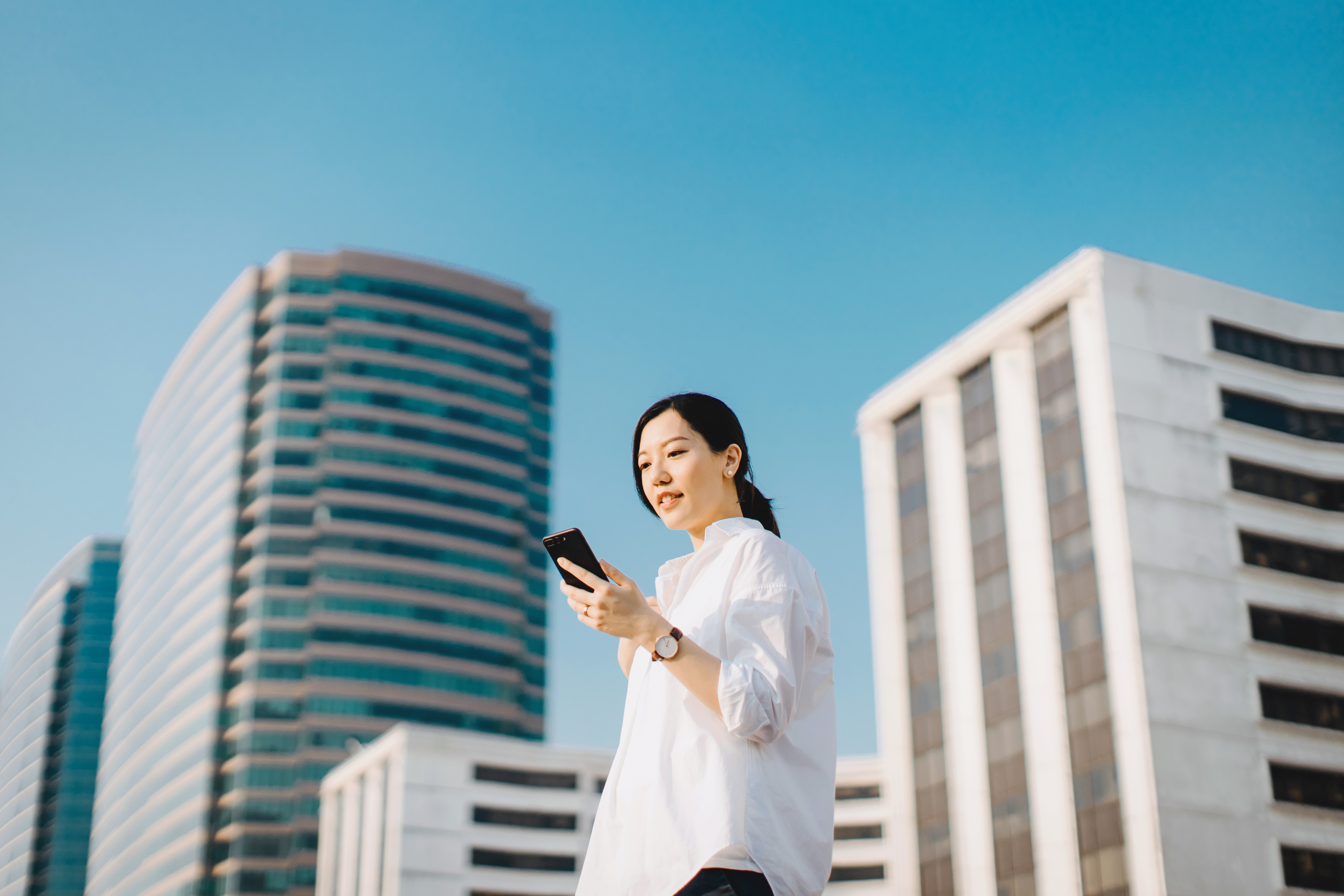 Confident young woman emailing on smartphone in city, against highrise city buildings and clear blue sky